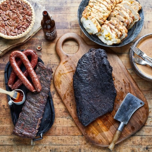 A wooden table displays smoked meats, including ribs and sausage, alongside bread, pie, and a bottle of sauce.