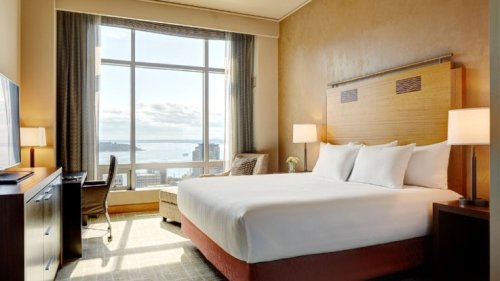 A modern hotel room with a large bed, desk, and a window showcasing a view of the city and water.
