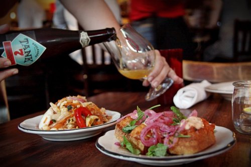 A hand pours a drink into a glass beside a plate of vibrant food, including a salad and a sandwich with toppings.