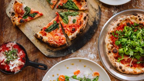 A wooden table with pizzas, one sliced, topped with basil and cheese, alongside a salad and a bowl of red sauce.