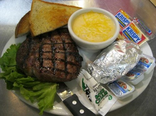 A plate with grilled steak, toast, a bowl of soup, a baked potato, and condiments.