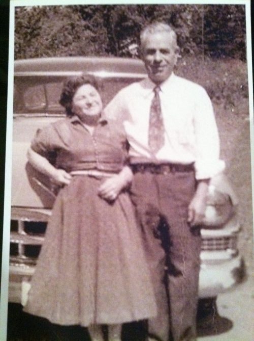 A vintage photo of a smiling couple standing beside an old car, with trees in the background.