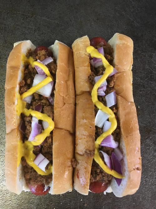 Two hot dogs in buns topped with ground meat, diced onions, and yellow mustard, placed on a metallic surface.