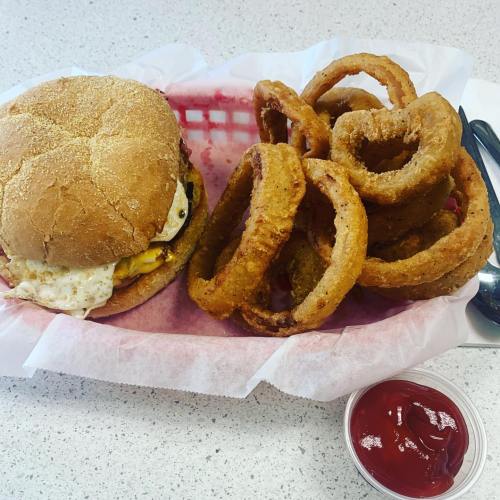 A burger with an egg on a bun, served with crispy onion rings and a side of ketchup in a takeout basket.