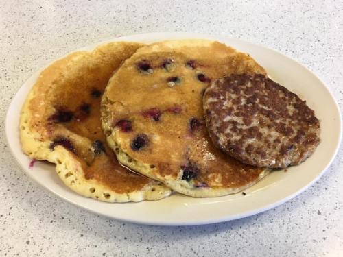 A plate with three pancakes, two with blueberries and syrup, and a sausage patty on the side.