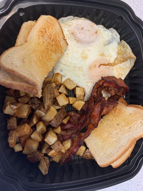 A takeout container with two fried eggs, crispy bacon, golden-brown potatoes, and two slices of toasted bread.