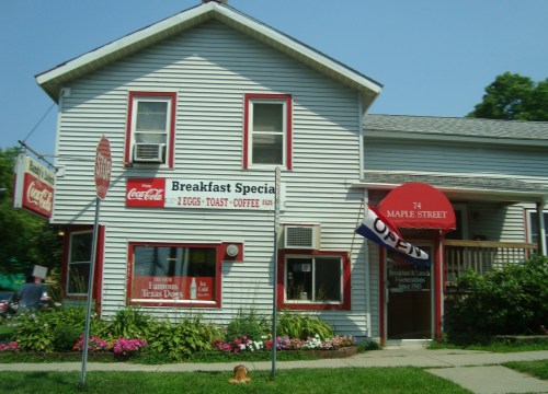 A quaint diner with a red awning, serving breakfast specials, surrounded by flowers and a "OPEN" sign.