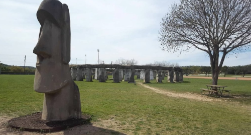 A large moai statue stands in a grassy area near a stone structure resembling Stonehenge, with a tree and picnic table nearby.