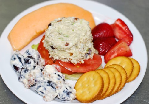 A plate with a mound of chicken salad, sliced tomatoes, strawberries, cantaloupe, and crackers.