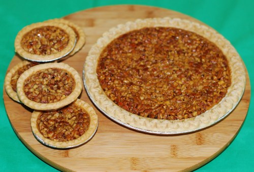 A large pecan pie alongside three smaller pecan tarts on a wooden cutting board, set against a green background.