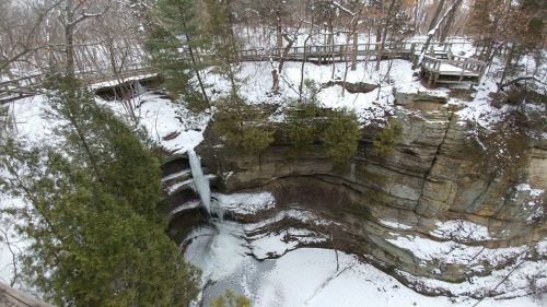 A snowy landscape featuring a waterfall cascading into a rocky gorge, surrounded by trees and a wooden walkway above.