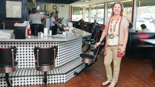 A woman stands smiling in a retro diner with black and white decor, while staff serve customers in the background.
