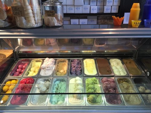 A display of various colorful gelato flavors in metal containers, with cones and cups in the background.