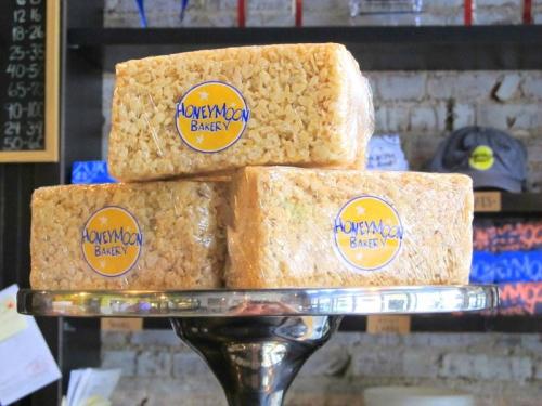 Three rectangular rice crispy treats wrapped in plastic, displayed on a silver cake stand with a HoneyMoon Bakery logo.