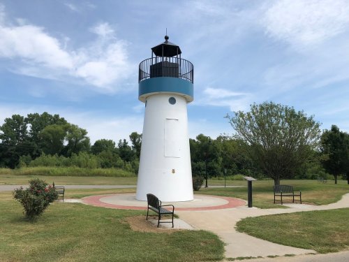 A white lighthouse structure surrounded by benches and greenery under a blue sky with scattered clouds.