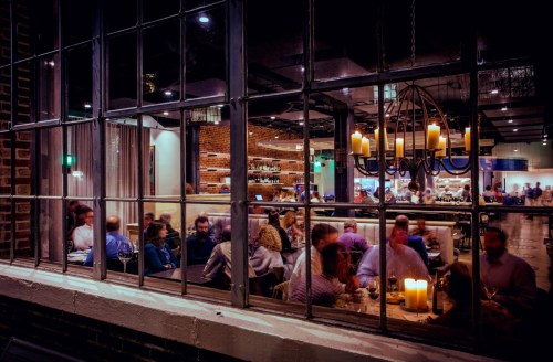 A dimly lit restaurant scene viewed through a large window, with diners and a chandelier visible inside.
