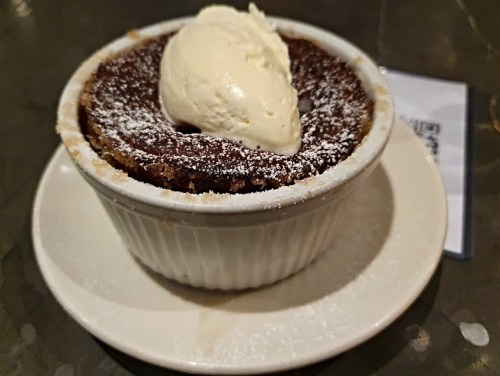 A chocolate dessert in a white ramekin, topped with a scoop of vanilla ice cream and dusted with powdered sugar.