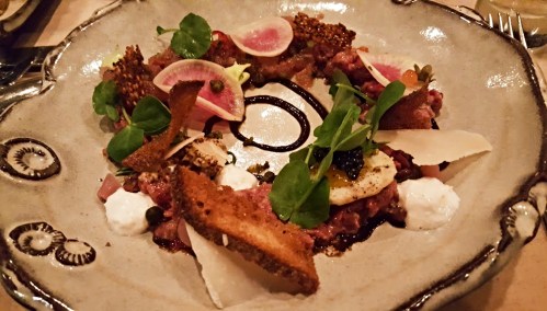 A gourmet dish featuring beef tartare, garnished with herbs, radish slices, crispy bread, and a drizzle of balsamic reduction.