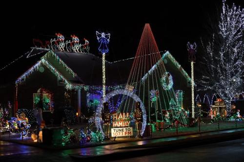 A festive house decorated with colorful Christmas lights, featuring a large tree, reindeer, and a sign to tune to 107.1 FM.