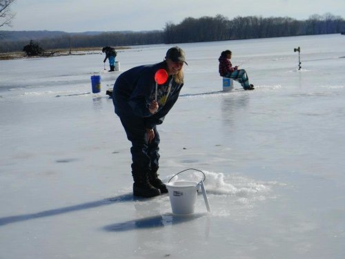 A person smiles while ice fishing on a frozen lake, with others sitting in the background. A bucket is nearby.