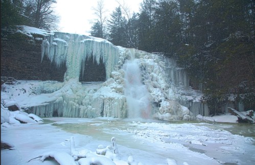A frozen waterfall surrounded by snow-covered rocks and trees, creating a serene winter landscape.
