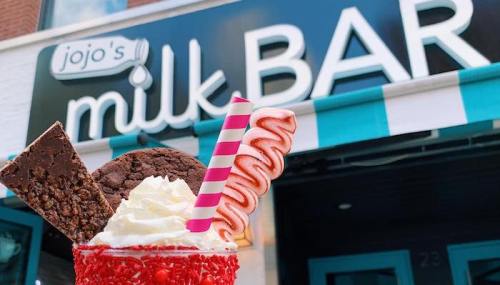 A colorful milkshake topped with whipped cream, a cookie, and a candy cane, in front of Jojo's Milk Bar sign.