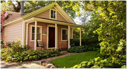 A charming pink cottage surrounded by greenery and flowers, featuring a welcoming front porch and a well-kept lawn.