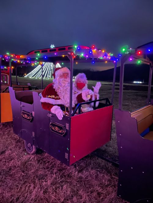 Santa and Mrs. Claus sit in a decorated train car, surrounded by colorful lights, with a festive backdrop.