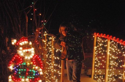 A child poses playfully next to a brightly lit Santa decoration amidst festive holiday lights at night.