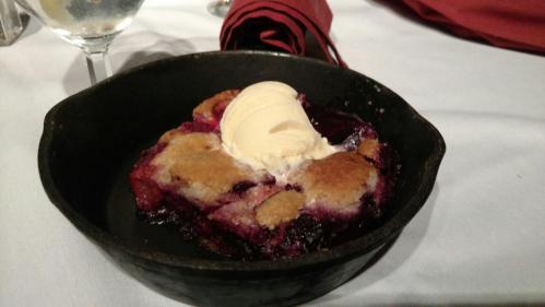A warm dessert in a cast iron skillet, featuring fruit filling topped with a scoop of vanilla ice cream.