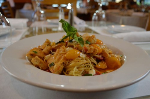 A plate of pasta topped with golden tomatoes, herbs, and crispy fried pieces, set in a restaurant setting.