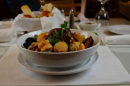 A bowl of mixed seafood, including shrimp, scallops, and mussels, garnished with herbs, served on a white tablecloth.