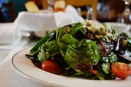 A fresh green salad with cherry tomatoes and crumbled cheese, served on a white plate with bread in the background.