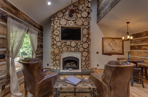Cozy living room with a log wall, fireplace, two armchairs, and a rustic wooden table, surrounded by greenery outside.