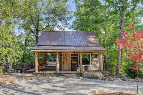 A rustic cabin with a metal roof, surrounded by trees and a gravel path, featuring a porch and colorful landscaping.