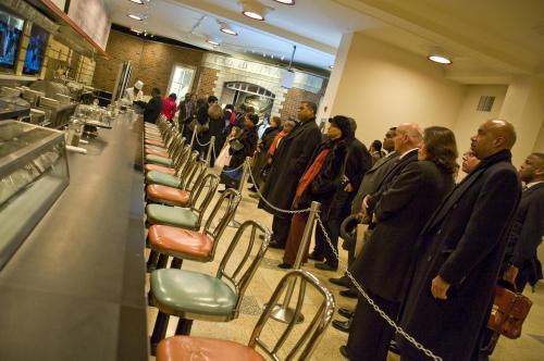 A line of people in formal attire waiting at a counter in a restaurant, with empty chairs and a chef in the background.