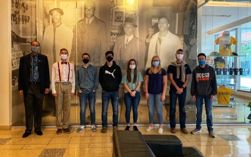 A group of eight people, wearing masks, stands in front of a historical black-and-white photo backdrop.