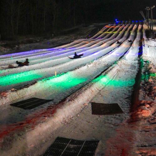 Colorful lights illuminate snow tubing lanes at night, with people sliding down the slopes.