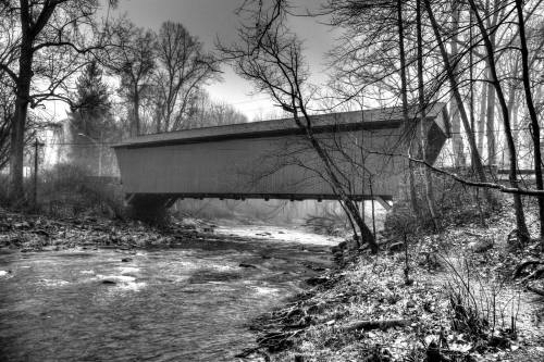 A black and white image of a covered bridge over a stream, surrounded by bare trees and a misty atmosphere.