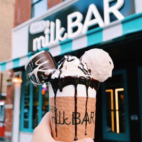 A hand holds a cup of ice cream topped with chocolate sauce and a chocolate treat, in front of a milk bar storefront.