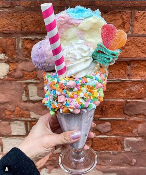 A colorful ice cream sundae topped with cotton candy, whipped cream, and a heart-shaped candy, served in a glass.