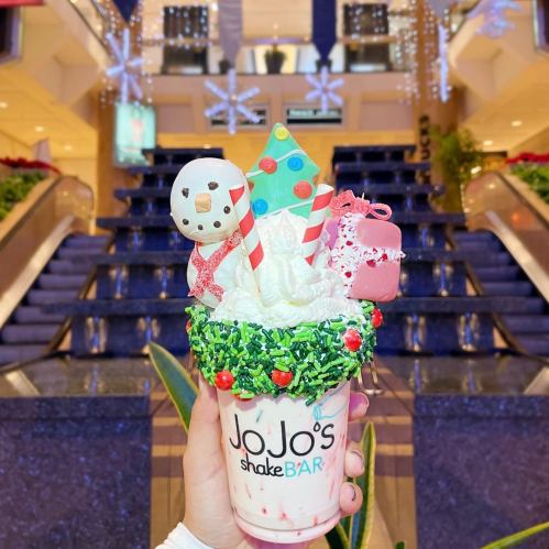 A festive milkshake topped with whipped cream, candy decorations, and a snowman cookie, held in front of a decorated staircase.