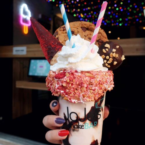 A colorful milkshake topped with whipped cream, a cookie, a chocolate-covered cone, and two straws, held in a hand.