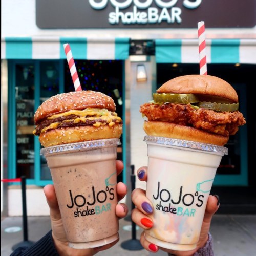 Two oversized milkshakes topped with burgers, one with a cheeseburger and the other with fried chicken, held in hands.