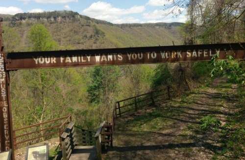 A rusted sign reading "Your family wants you to work safely" above a wooded path with a scenic view in the background.