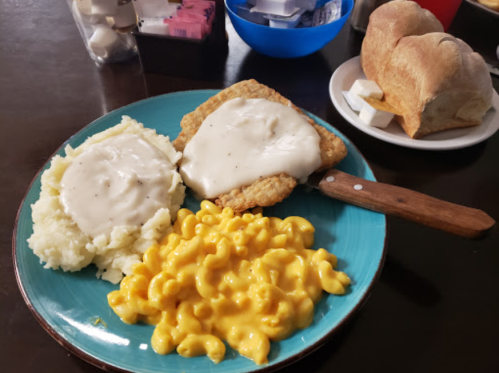 A plate with fried chicken covered in gravy, mashed potatoes, macaroni and cheese, and rolls in the background.