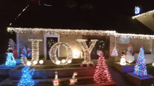 A festive house decorated with bright lights, featuring the word "JOY" and colorful Christmas trees and decorations.