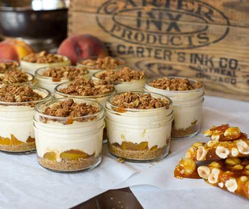 Dessert jars with layers of creamy filling, peaches, and crumbled topping, alongside a piece of nut brittle.