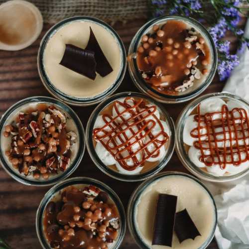 A variety of desserts in glass jars, topped with chocolate curls, caramel, nuts, and crunchy toppings.