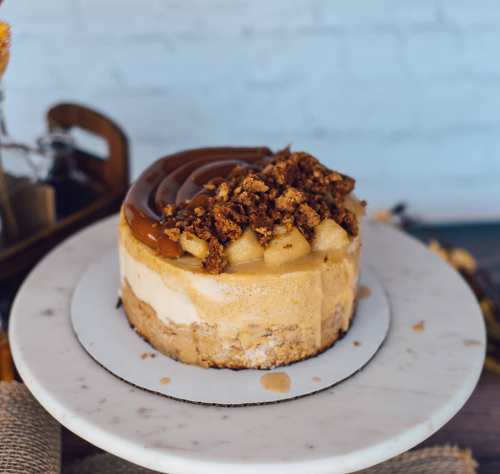 A delicious layered cheesecake topped with caramel, crumbled cookies, and pieces of fruit on a marble cake stand.